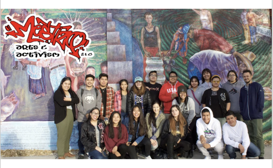 A group photo of young adults and mentors from the MESTIZX Arts & Activism SLC collective, standing in front of a vivid community mural. The mural depicts agricultural and cultural imagery, with the group smiling and posed together, suggesting solidarity, grassroots organizing, and intergenerational cultural preservation. The “MESTIZX Arts & Activism SLC” logo is spray-paint styled in the upper left.